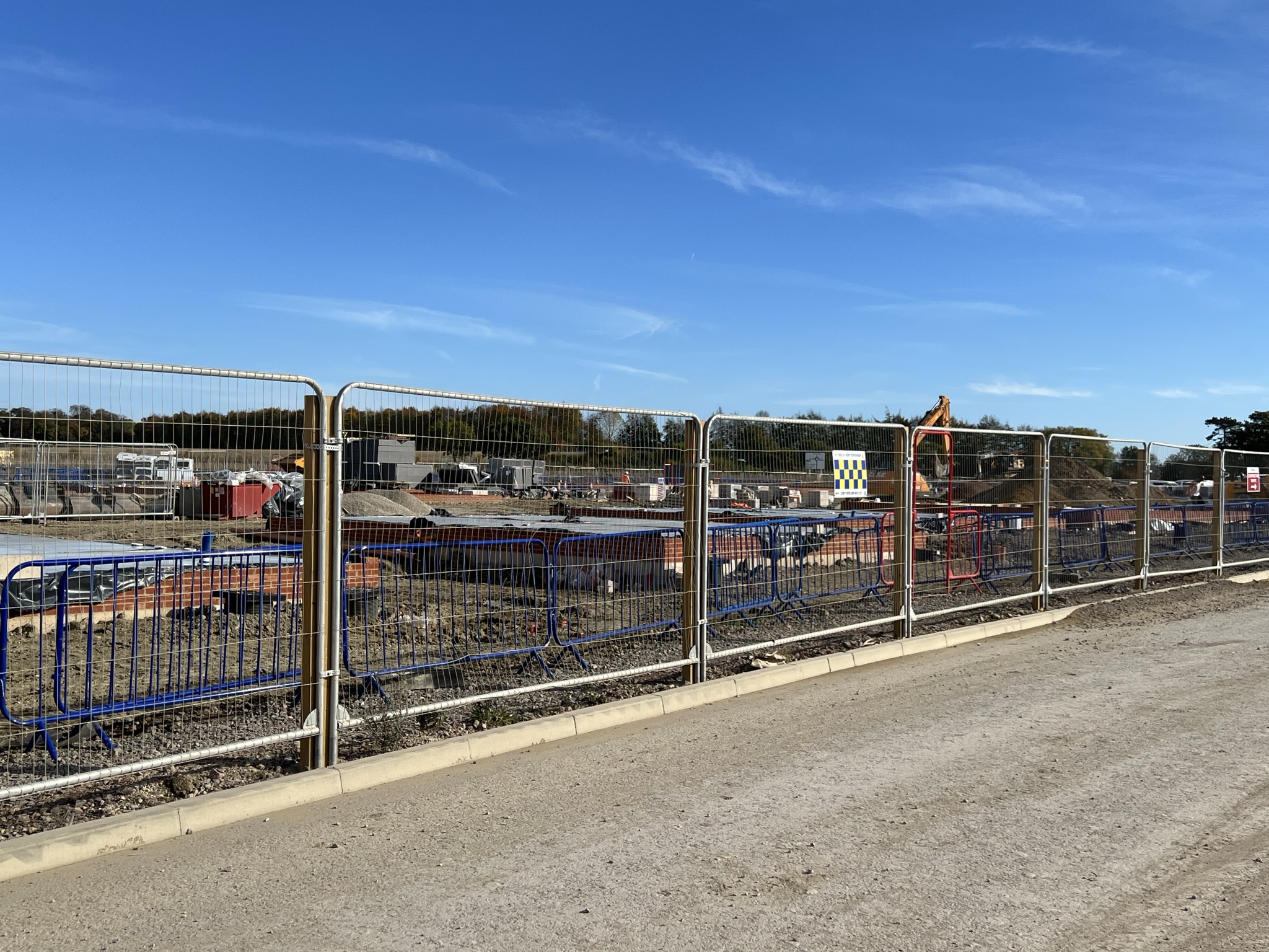 Curving post-supported Heras mesh fencing with blue pedestrian barriers alongside an active construction site under a clear blue sky